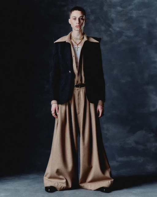 Full-length studio shot of a person wearing a black jacket, beige shirt, tan pants, jewelry, and black shoes against a dark backdrop.