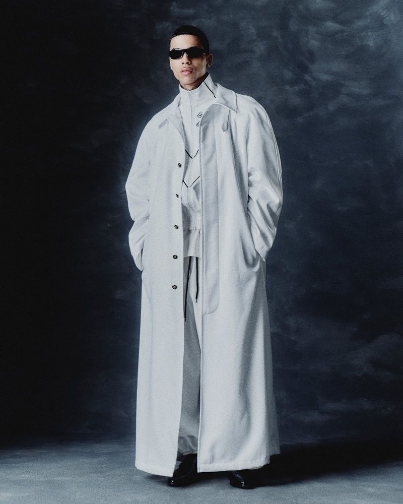 A full-length shot of a male model wearing sunglasses and a long white coat in a studio setting with a muted grey background.
