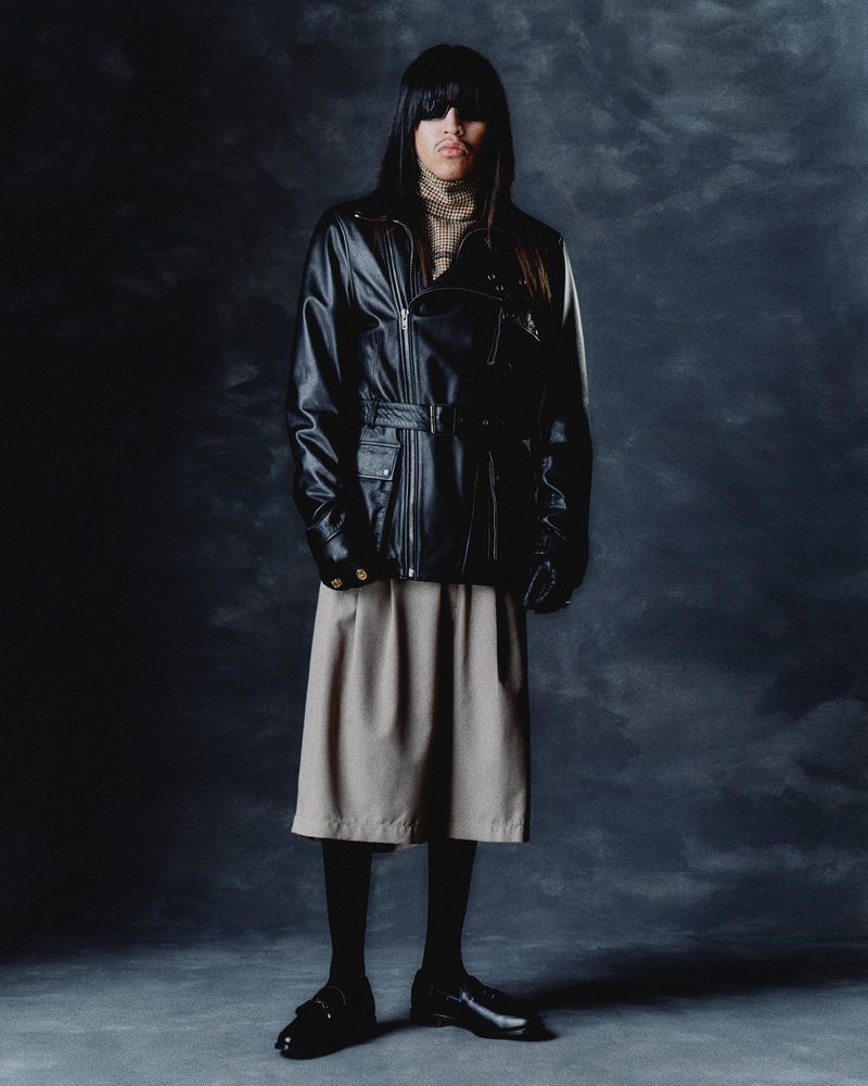 A person in a black leather motorcycle jacket, beige skirt, black tights and loafers stands in front of a mottled grey background.