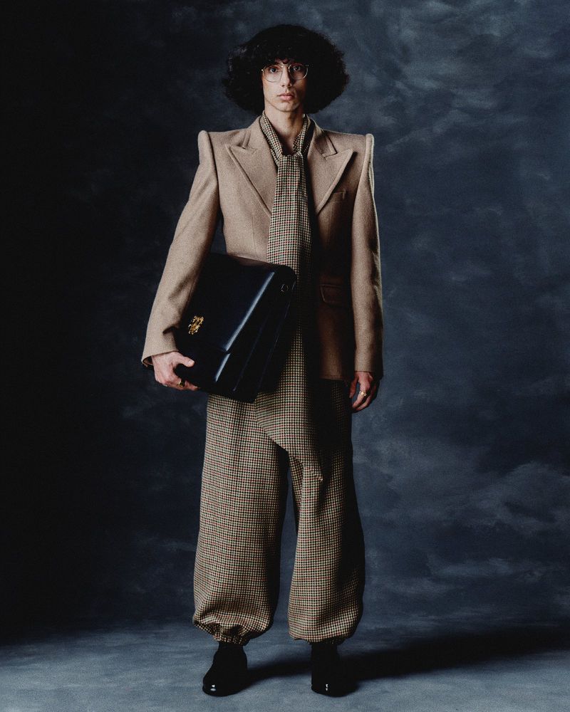 Male model in a brown suit and tie, holding a black briefcase, poses against a dark, textured background in a studio shot.