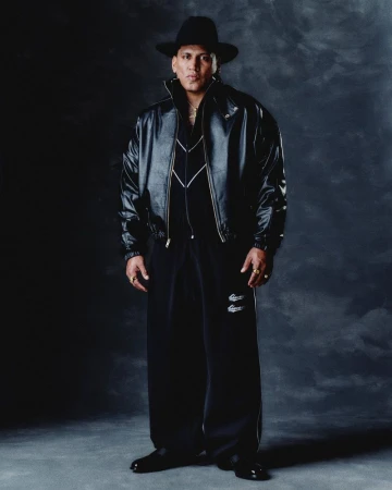 Man in an oversized black leather jacket, wide-leg track pants, and a wide-brim black fedora against a dark textured backdrop.