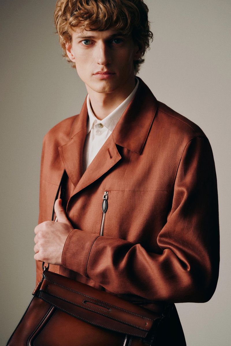 Portrait of a blond-haired male model in a rust-colored jacket holding a brown leather satchel against a light grey studio backdrop.