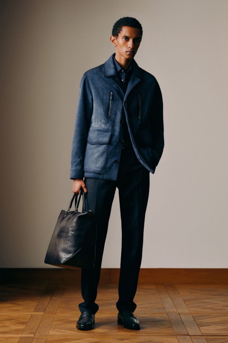 A dark-haired male fashion model poses indoors in a blue jacket and black pants, holding a large black leather bag with a wood floor.