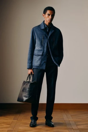 A dark-haired male fashion model poses indoors in a blue jacket and black pants, holding a large black leather bag with a wood floor.