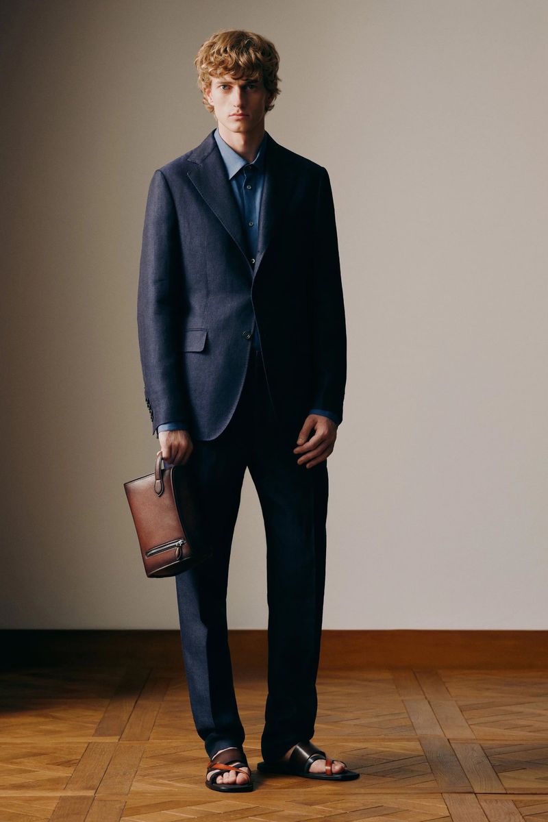 Fashionable adult male posing in a navy blue suit with a brown leather bag and sandals, standing on a wood parquet floor.