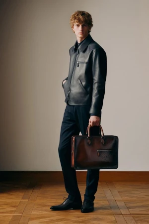 Man in black leather jacket and trousers holding a brown leather briefcase, standing on a parquet floor with a neutral wall background.