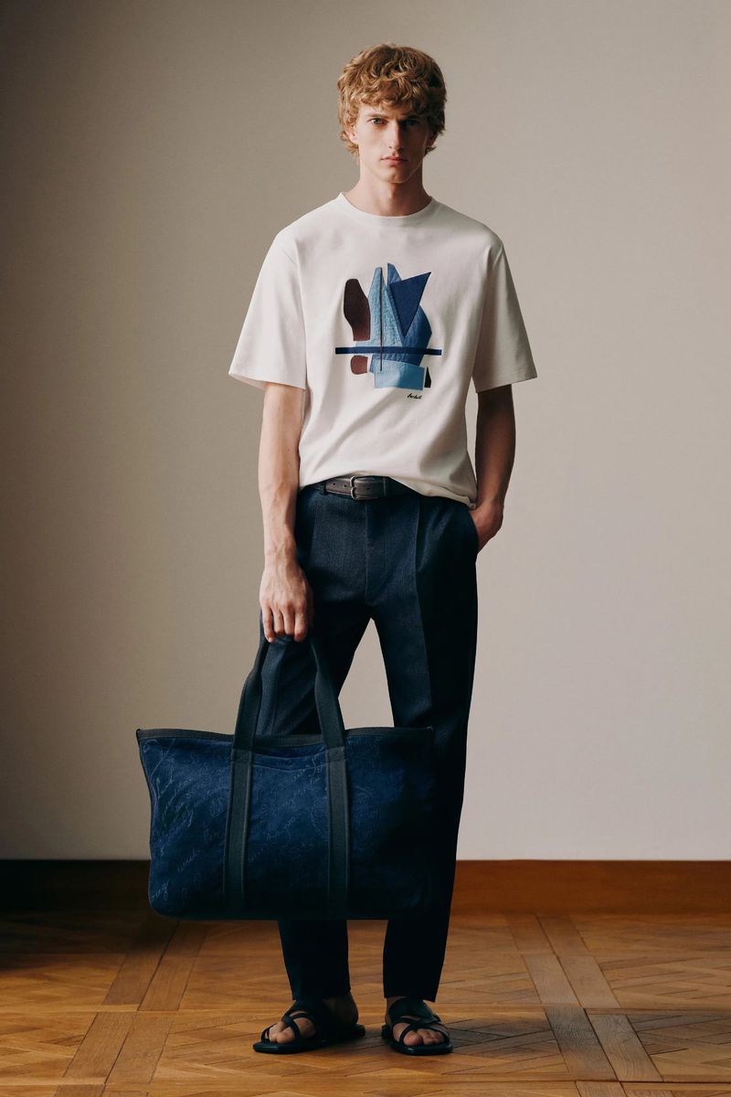 Male fashion model poses indoors with t-shirt with abstract art, dark trousers, leather sandals, and blue textured duffel bag.