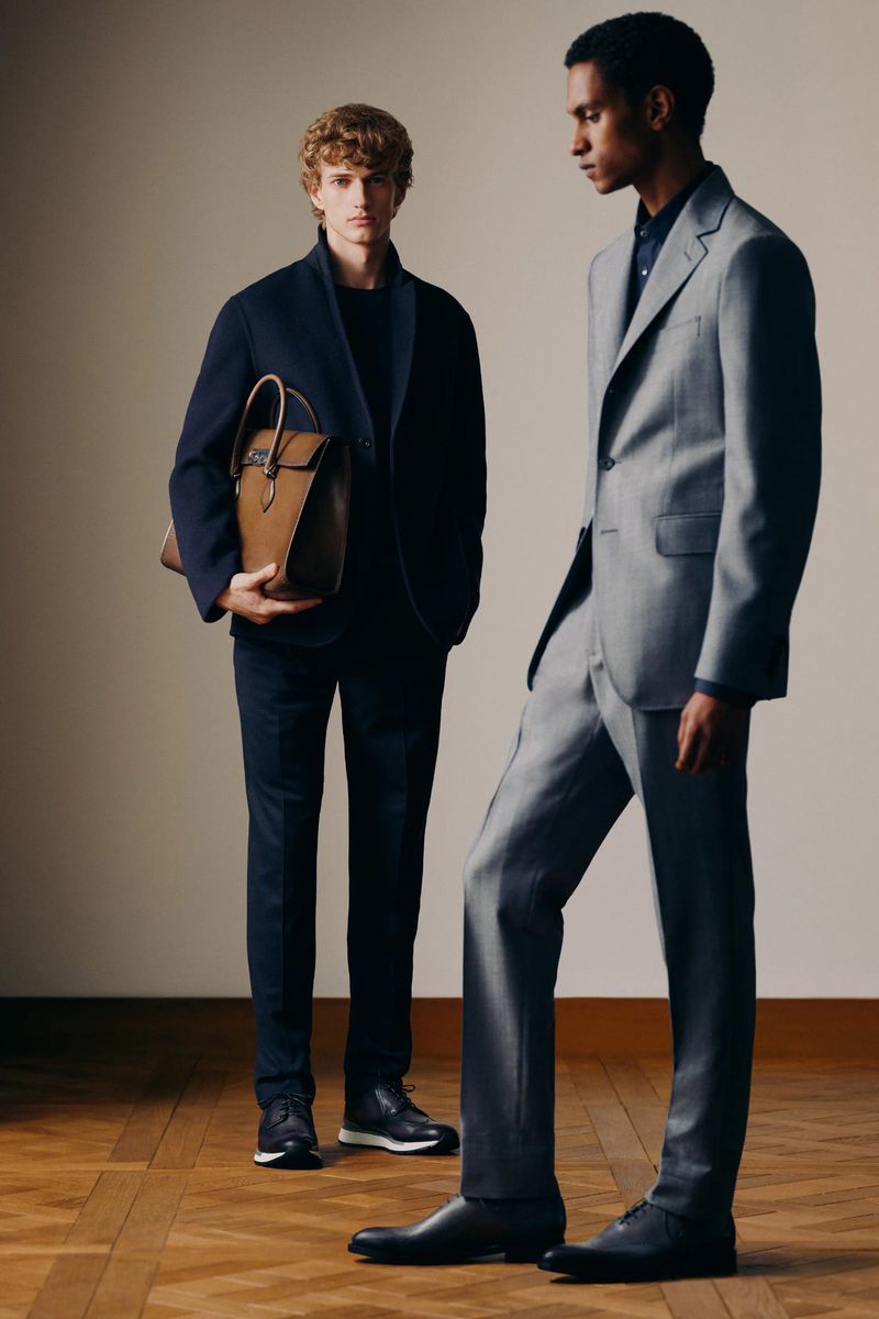 Two men in suits stand indoors. One holds a leather satchel. The men are wearing formal suits in a studio setting.