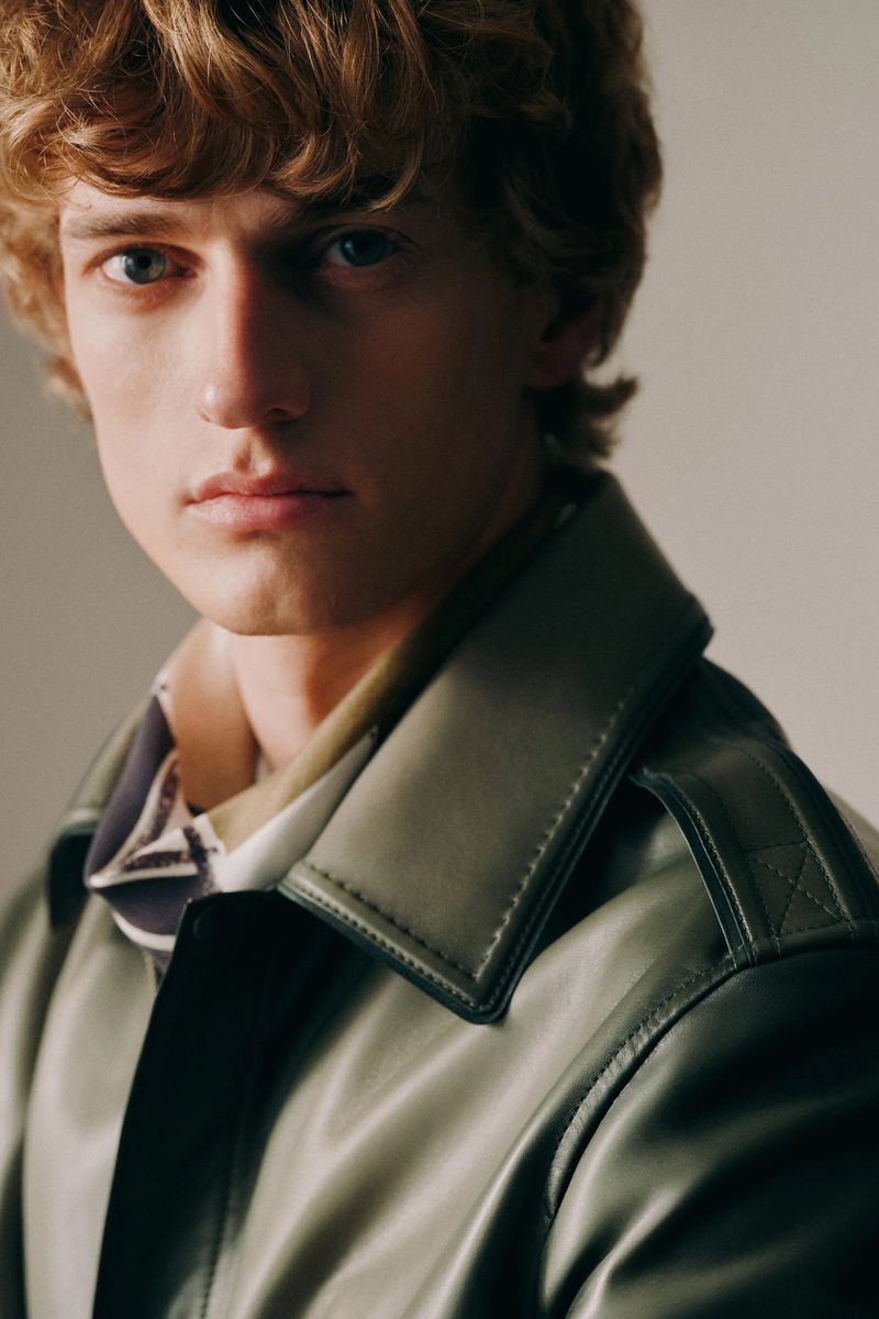 Close-up portrait of a young adult male with wavy blonde hair wearing an olive-green trench coat against a neutral background.