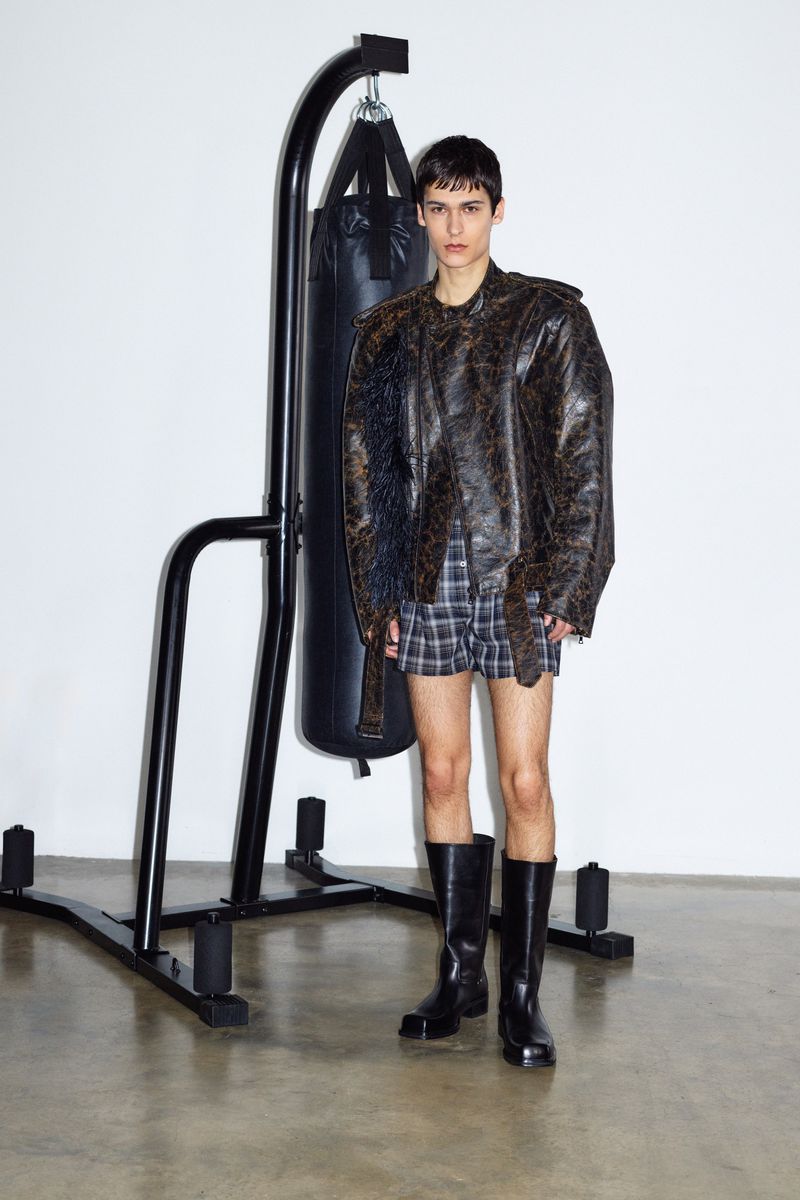 Fashionable adult stands near a black punching bag, wearing a snakeskin jacket, plaid shorts, black boots in a studio environment.