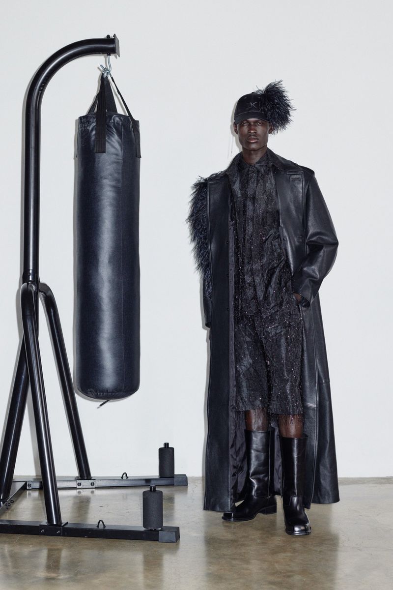 Dark-skinned male model in black leather trench coat, textured top and shorts next to black punching bag and metal stand in studio.