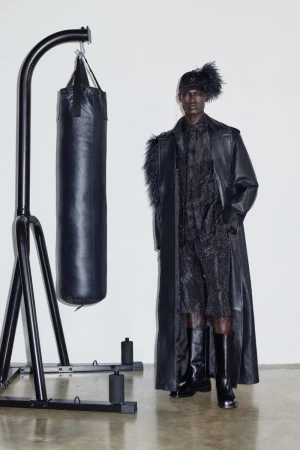 Dark-skinned male model in black leather trench coat, textured top and shorts next to black punching bag and metal stand in studio.