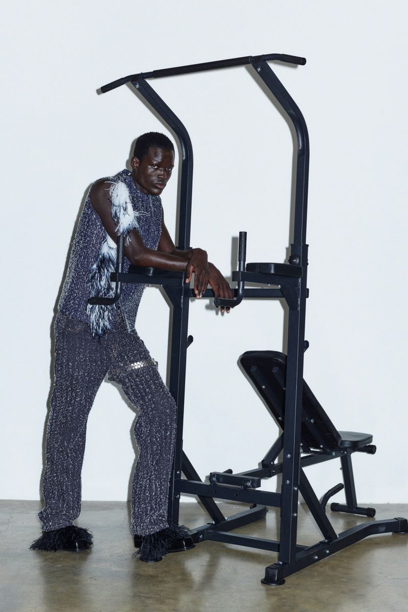 Athletic, Black adult male posing next to black exercise pull-up machine, wearing textured top and pants, with a white background.