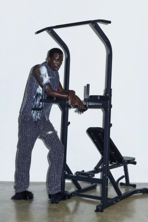 Athletic, Black adult male posing next to black exercise pull-up machine, wearing textured top and pants, with a white background.