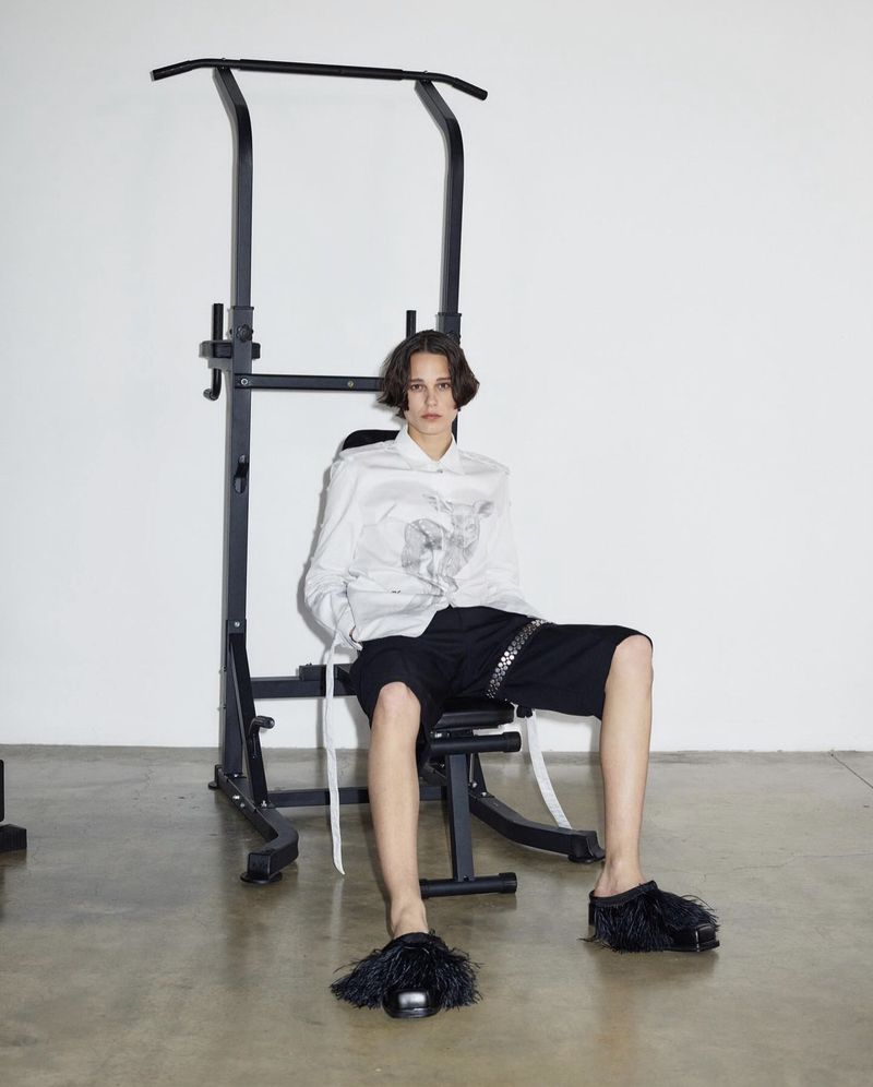 Fashion shot of a model in a white shirt and black shorts sitting on a black weight bench in a bright white studio with concrete floors.