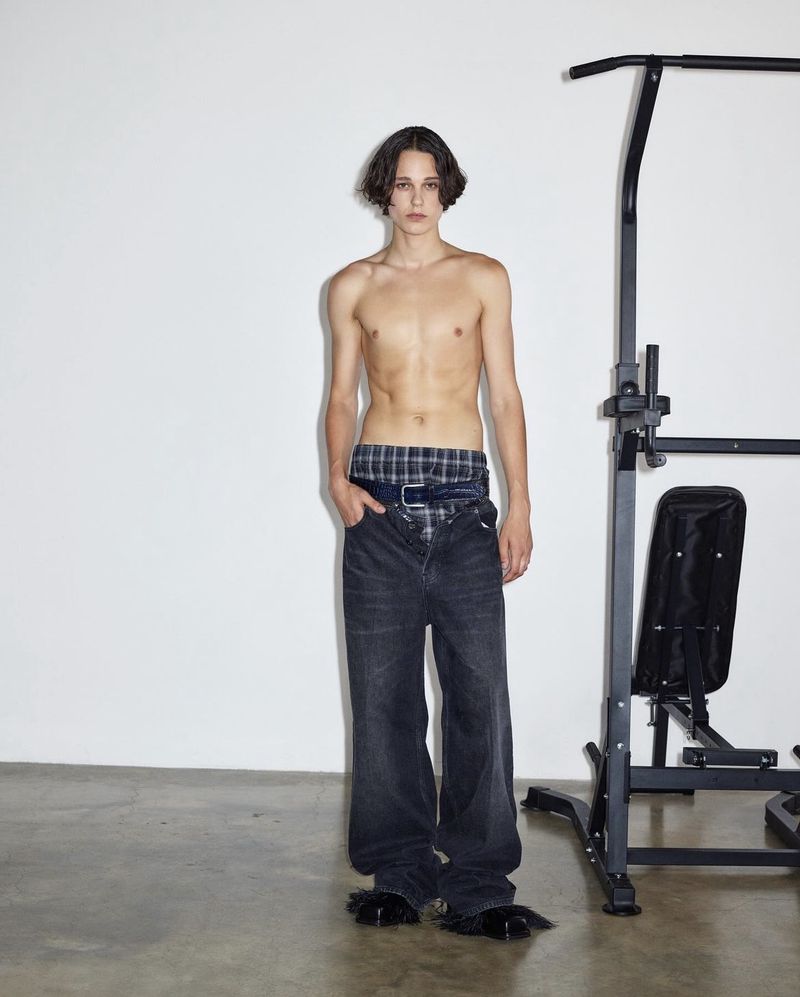Shirtless adult male in baggy denim stands indoors near a black metal pull-up bar against a white wall. Fashion editorial photograph.