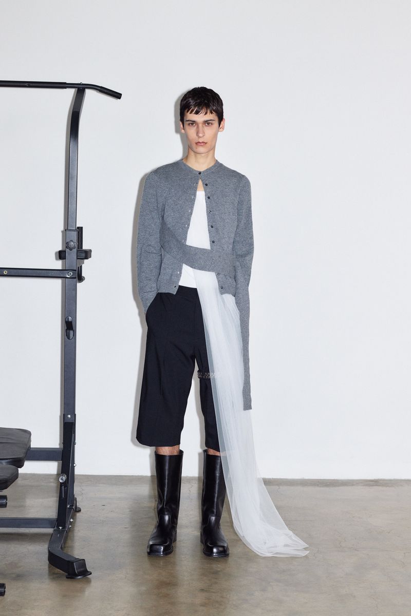 Fashion model posing indoors near a pull-up bar, wearing a grey cardigan, white top, black shorts, and black boots in a studio setting.