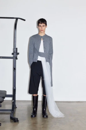 Fashion model posing indoors near a pull-up bar, wearing a grey cardigan, white top, black shorts, and black boots in a studio setting.