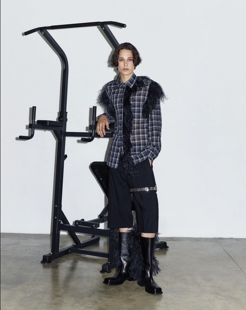 Woman poses in a plaid shirt with feathered trim and black culottes alongside a black metal pull-up bar in a photo studio.