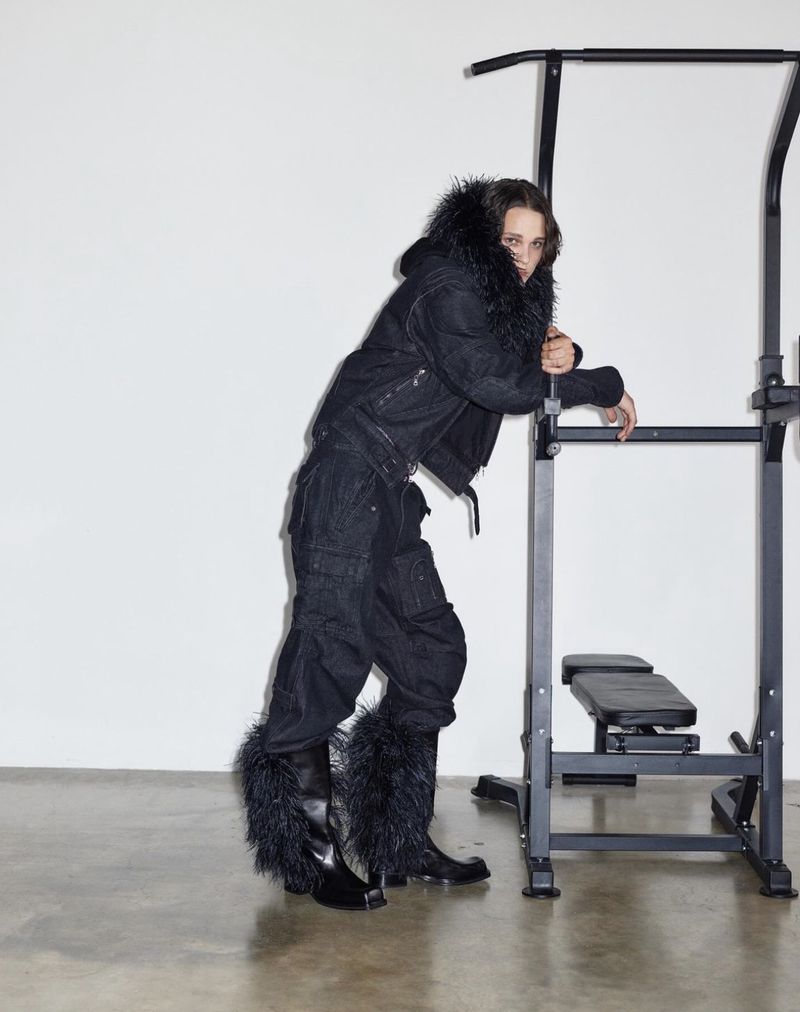 Person in black fur-trimmed outfit leaning against a black metal pull-up bar indoors, on a concrete floor, with a white wall background.