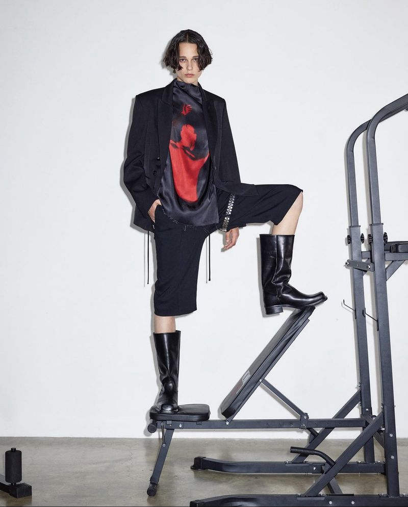 Person in a black suit and boots posing on fitness equipment against a white wall, creating a high-fashion editorial shot.
