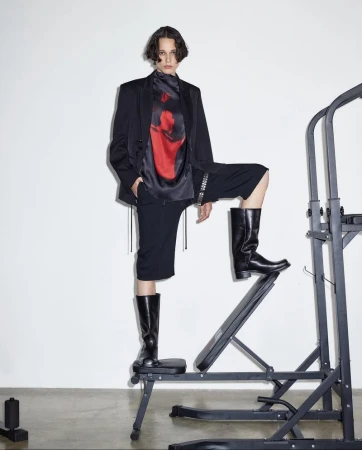 Person in a black suit and boots posing on fitness equipment against a white wall, creating a high-fashion editorial shot.