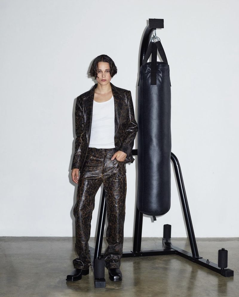 Woman in a brown patterned suit stands next to a black punching bag in a white photo studio, posing in a confident stance.