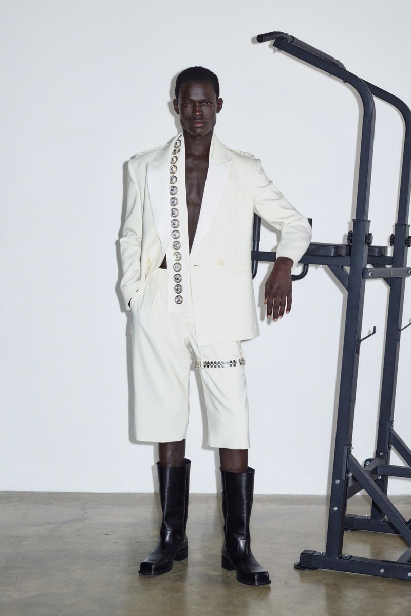 Dark-skinned adult male fashion model poses indoors wearing an off-white suit with black boots and a weight rack in the background.