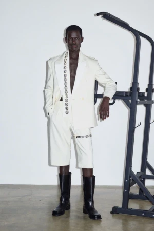 Dark-skinned adult male fashion model poses indoors wearing an off-white suit with black boots and a weight rack in the background.