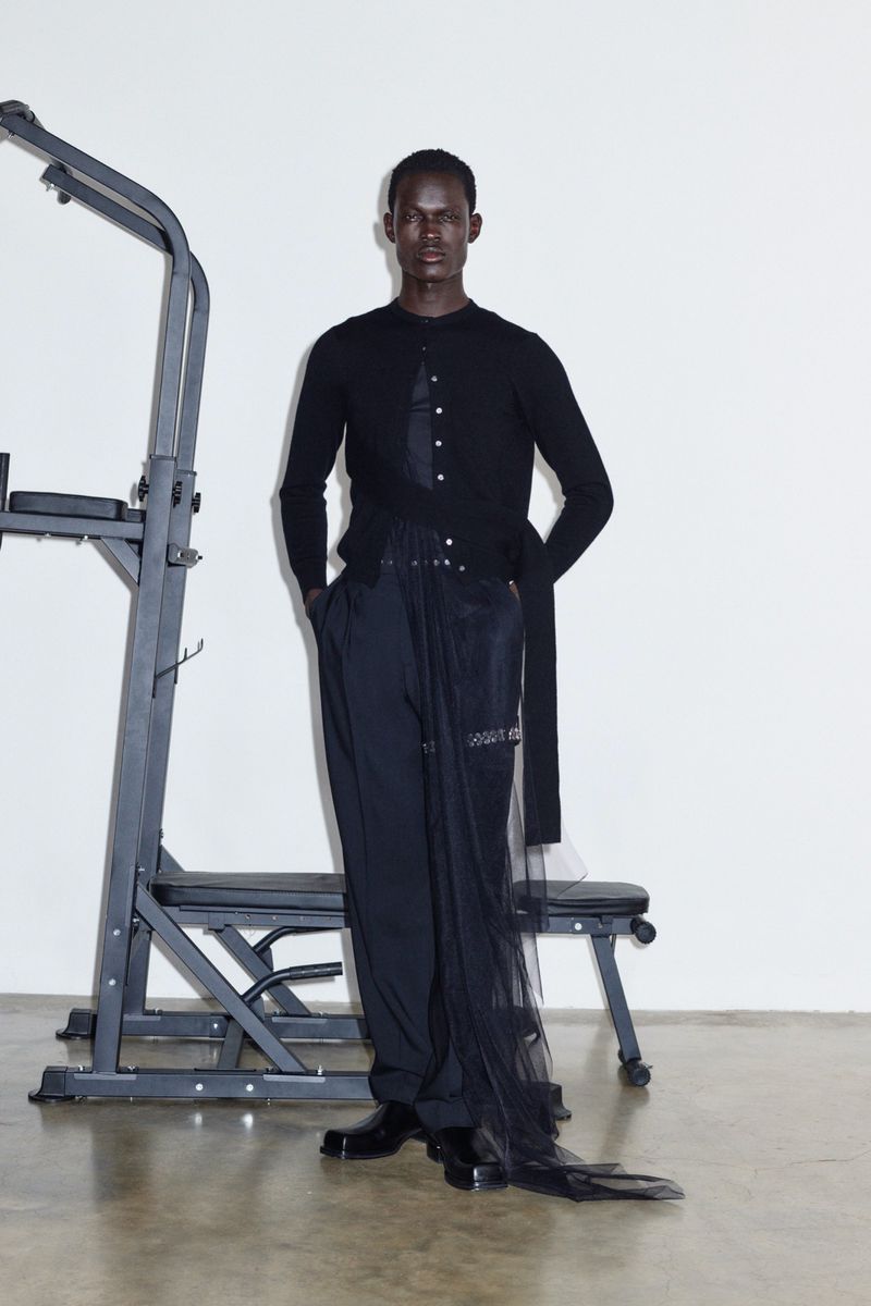 An adult male poses indoors, wearing a black cardigan, dark pants with sheer black fabric, and black shoes, standing next to gym equipment.