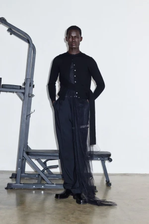An adult male poses indoors, wearing a black cardigan, dark pants with sheer black fabric, and black shoes, standing next to gym equipment.