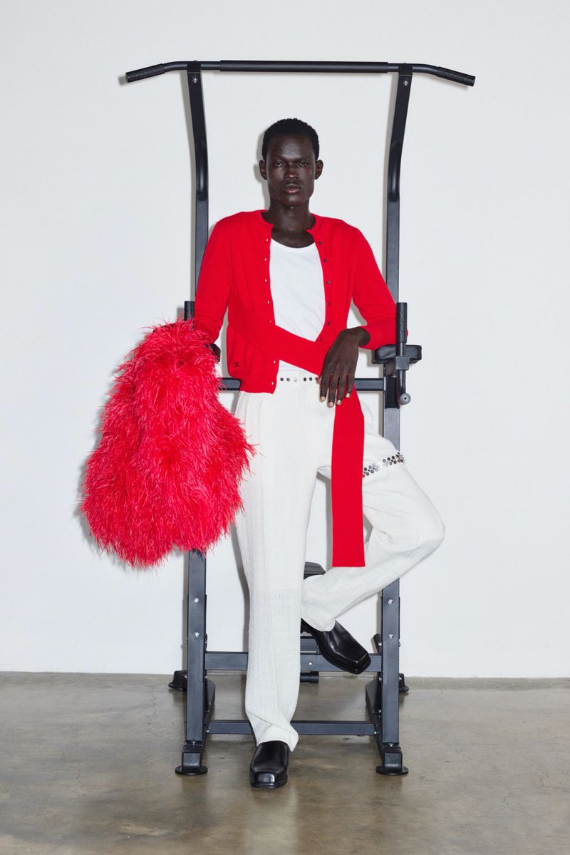A slender Black male fashion model poses indoors in casual wear, leaning on a black metal pull-up bar with red feathery coat.