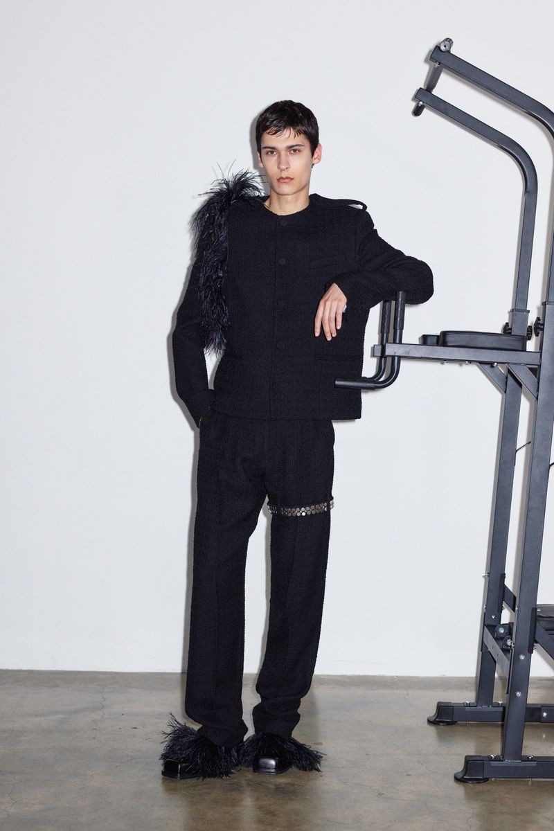 Fashion portrait of a young adult male model in a dark suit with feather details, posing next to a metal pull-up bar against a white wall.