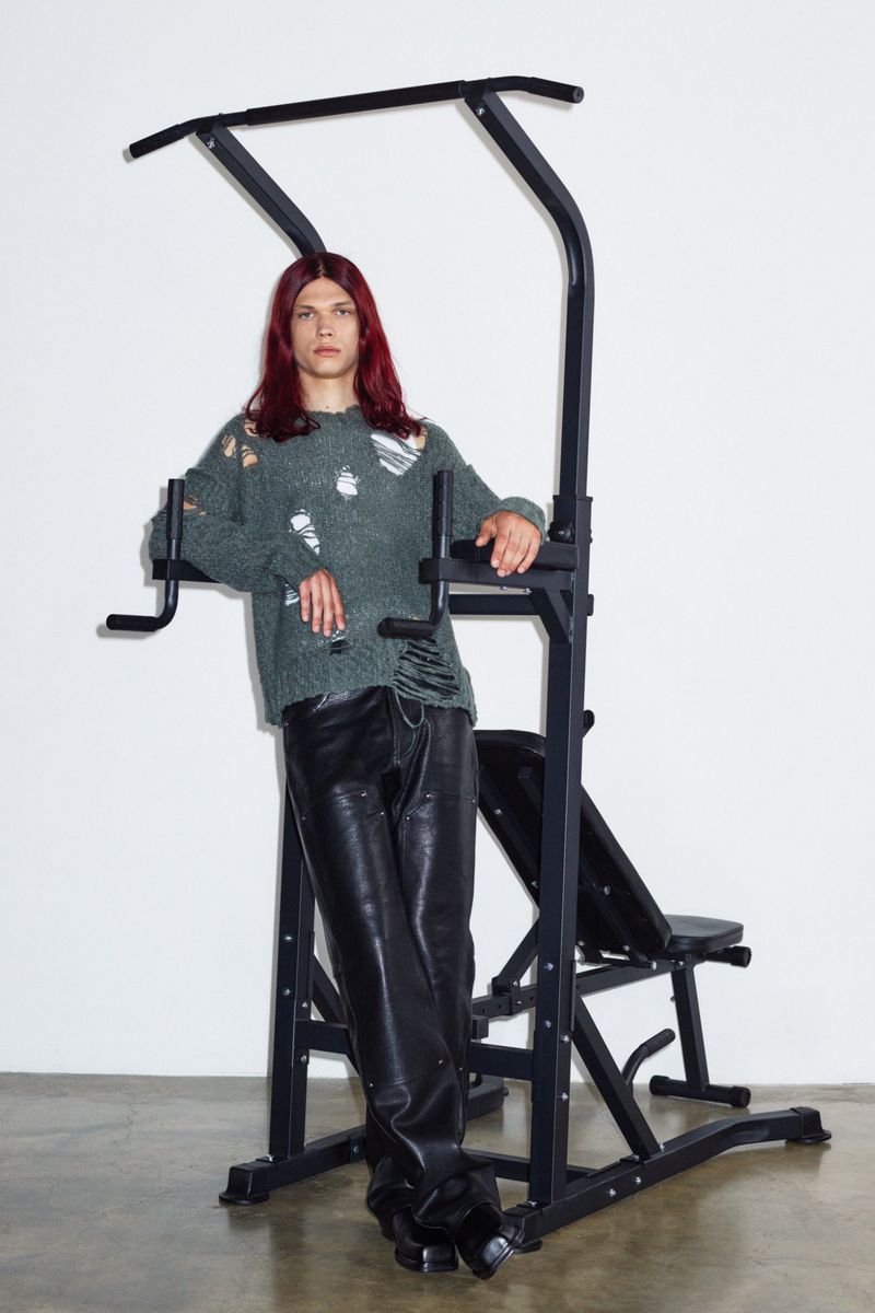 Man with long brown hair in distressed green sweater and black leather pants posing by black workout equipment against a white background.