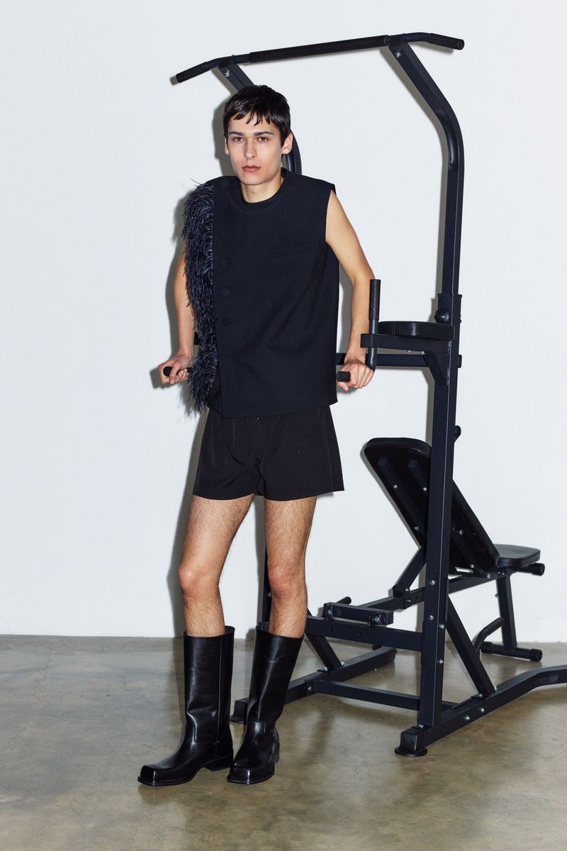 Person poses in black sleeveless top and shorts near exercise machine, wearing tall leather boots against a white background.
