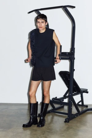 Person poses in black sleeveless top and shorts near exercise machine, wearing tall leather boots against a white background.