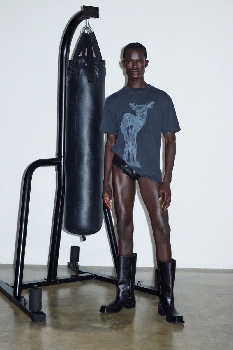 Dark-skinned male fashion model poses in black t-shirt, briefs, and boots next to black punching bag, in a white photo studio.