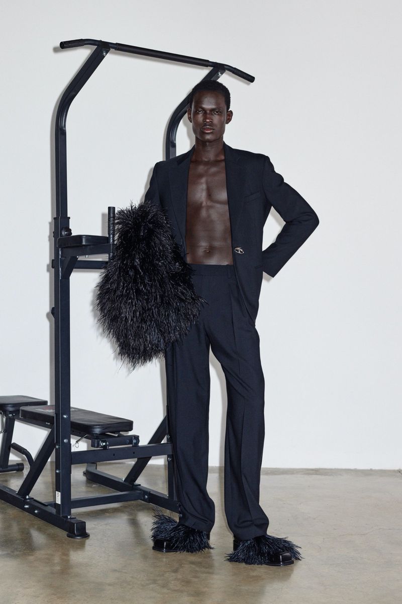 Man poses in black suit and furry slippers next to workout equipment in a studio with a white wall and concrete floor. Fashion editorial.