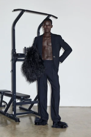 Man poses in black suit and furry slippers next to workout equipment in a studio with a white wall and concrete floor. Fashion editorial.