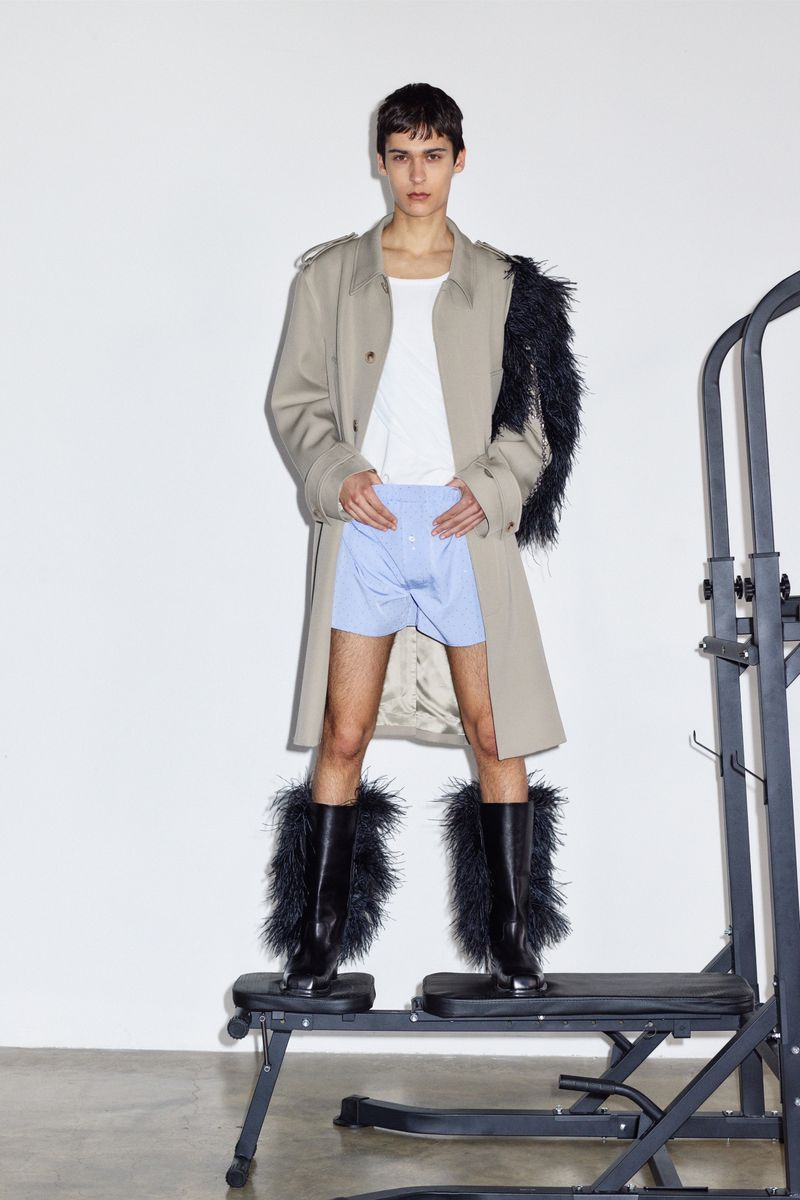 Fashion image of a slender adult male in a tan trench coat, blue shorts, and black feather boots on a platform in a studio.