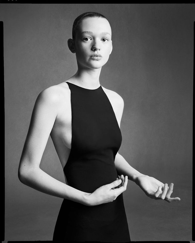 Studio shot of a woman with a closely shaved head, wearing a black dress with a high neckline against a grey backdrop with soft lighting.