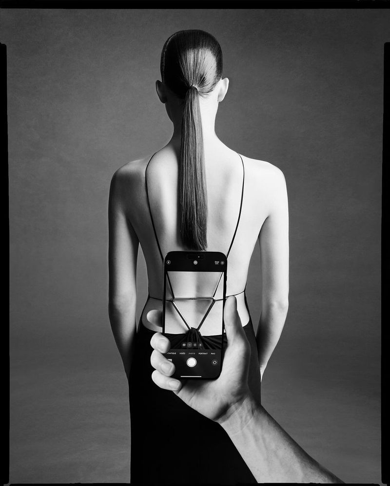 Back view of a woman in a black dress with a long ponytail, a hand holds up a smartphone in front of the dress's straps.