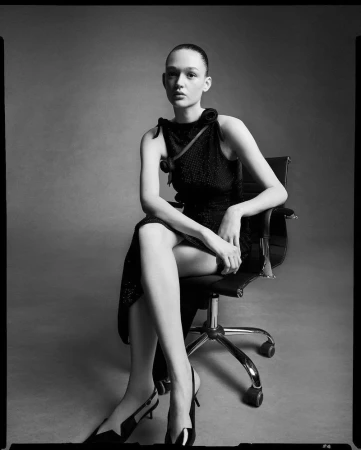 Monochrome portrait of a model in a dark dress seated on a black office chair against a gray background in a photo studio.