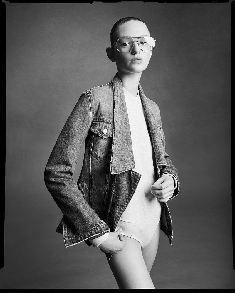 Black and white portrait of a young woman in glasses and a denim jacket against a grey backdrop in a fashion studio setting.
