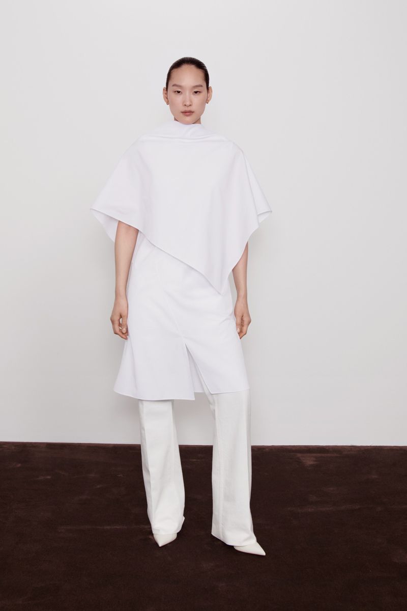 Asian woman in a white, asymmetrical tunic top and wide-legged pants posing in a white photo studio against a solid white backdrop.