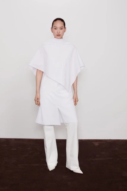 Asian woman in a white, asymmetrical tunic top and wide-legged pants posing in a white photo studio against a solid white backdrop.