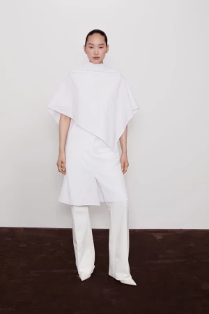 Asian woman in a white, asymmetrical tunic top and wide-legged pants posing in a white photo studio against a solid white backdrop.