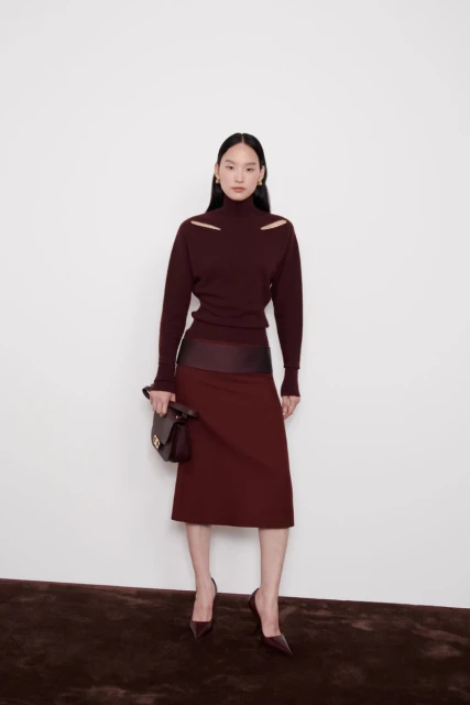 A slender Asian woman poses in a studio, wearing a burgundy turtleneck sweater with shoulder cutouts, a matching skirt, and heels.