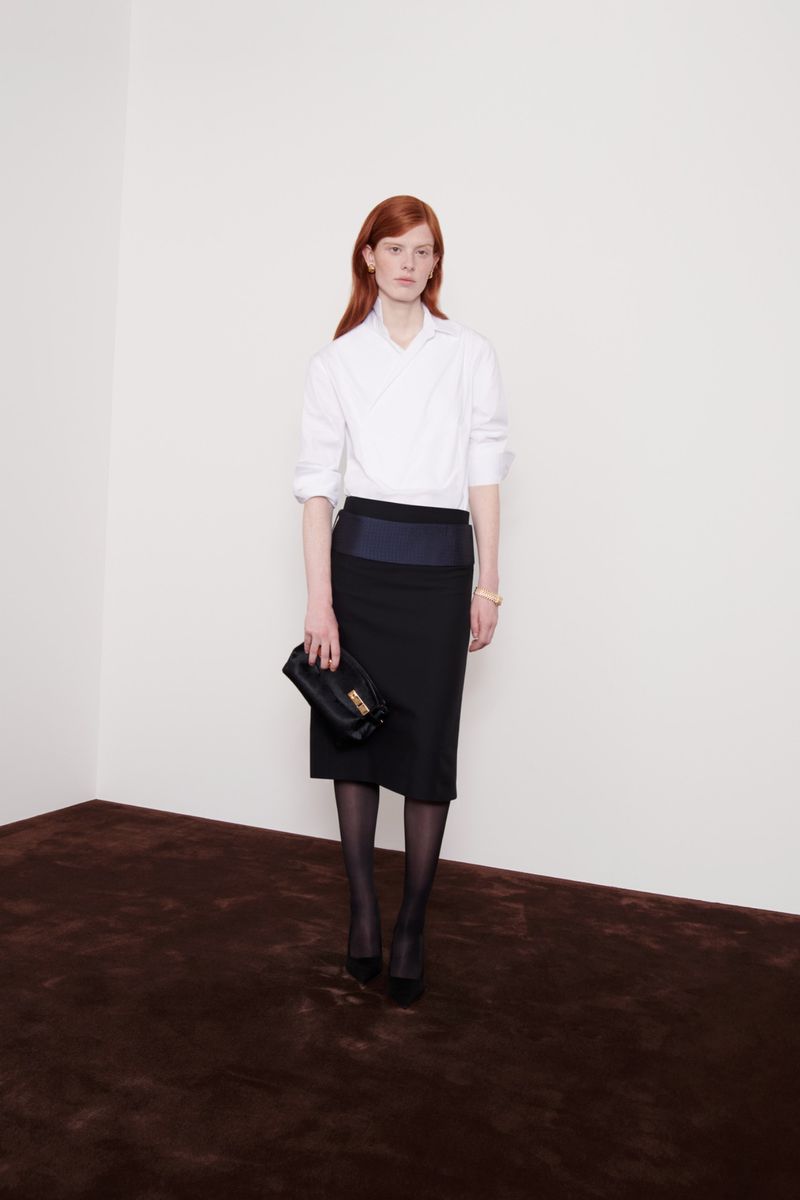 A red-haired woman in a white blouse and black skirt poses indoors with a dark handbag, looking straight at the camera in a fashion style.