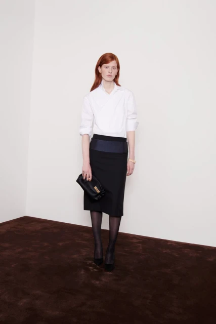 A red-haired woman in a white blouse and black skirt poses indoors with a dark handbag, looking straight at the camera in a fashion style.